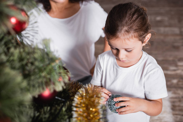 Selective focus of girl standing near mother and holding bauble near pine  - Φωτογραφία, εικόνα