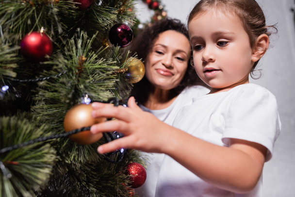 Low angle view of girl touching christmas ball on pine near mother at home - Photo, Image