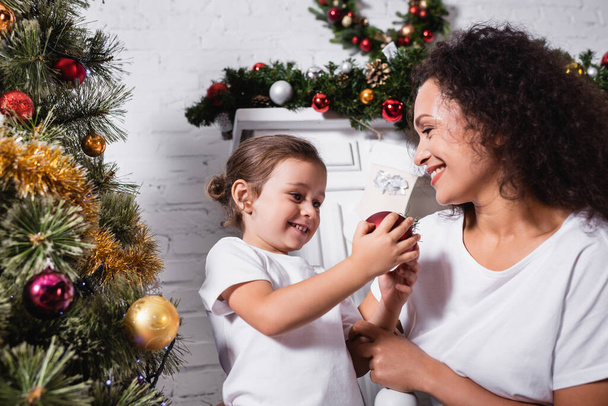 Mère et fille avec boule de Noël près de pin décoré à la maison - Photo, image