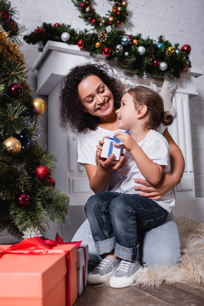 Mother embracing daughter with gift box near decorated pine and fireplace at home - Photo, Image