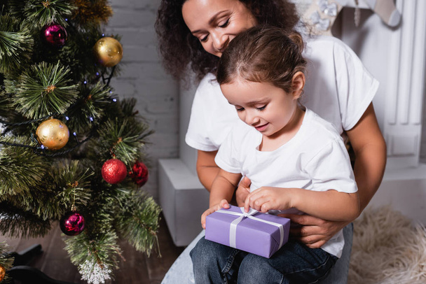 Madre con hija abriendo caja de regalo cerca de pino festivo en casa - Foto, Imagen