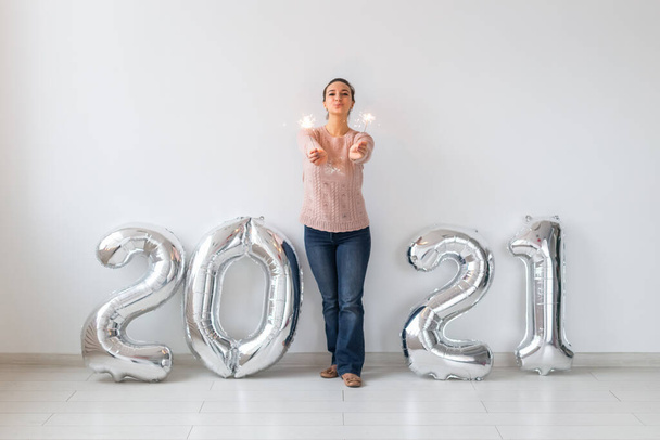 Πρωτοχρονιά γιορτή και κόμμα έννοια - Ευτυχισμένη νεαρή γυναίκα με sparklers κοντά σε ασημένια μπαλόνια 2021 σε λευκό φόντο. - Φωτογραφία, εικόνα
