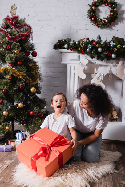 Chica con la boca abierta sosteniendo gran caja de regalo cerca de la madre y el pino festivo en casa - Foto, Imagen