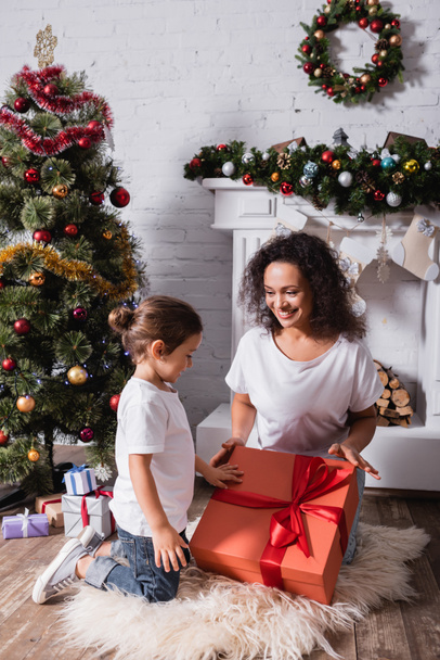 Mother with daughter touching big gift box near fireplace and pine at home - Foto, Imagem