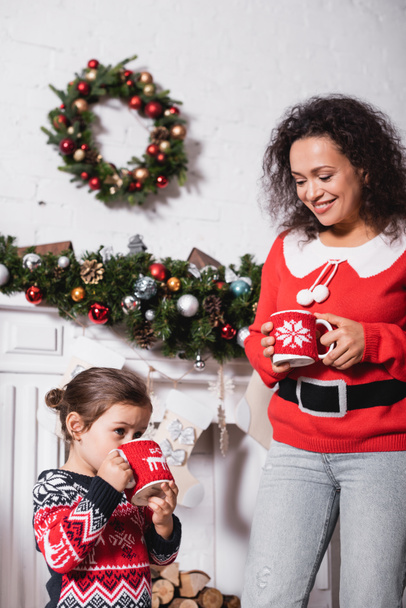 Little girl drinking from cup, standing with mother near decorated fireplace - Photo, Image