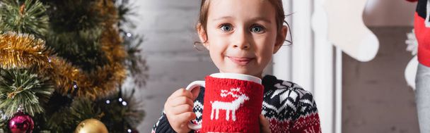 Panoramic shot of girl holding cup with knitted holder and looking at camera - Фото, зображення