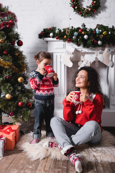 Mother sitting on rug near daughter drinking from cup near festive pine at home - Zdjęcie, obraz