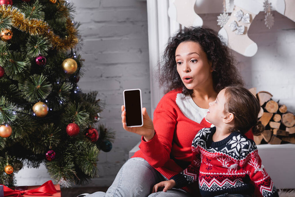 Fille avec mère montrant téléphone portable avec écran blanc près de pin et cheminée - Photo, image