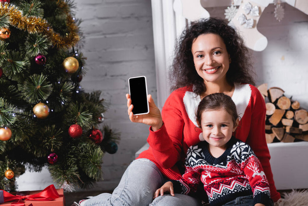Filha e mãe mostrando smartphone com tela em branco perto de pinho decorado - Foto, Imagem