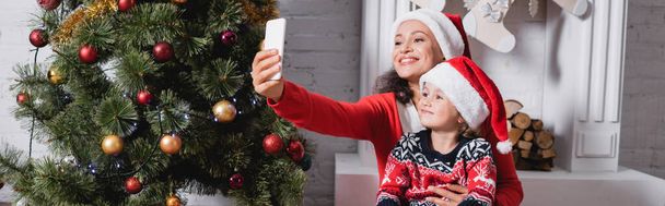 Panoramic shot of mother and daughter in santa hats taking selfie near pine - Photo, Image