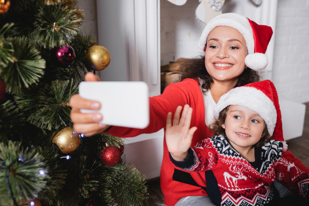 Mother and daughter with waving hand taking selfie near decorated pine - Foto, imagen