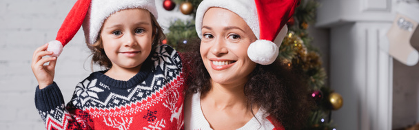 Panoramic shot of mother and daughter in santa hats looking at camera - Fotoğraf, Görsel
