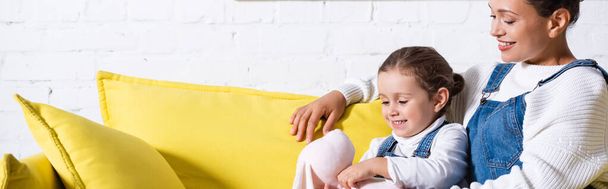 Πανοραμική φωτογραφία της μαμάς και της κόρης με μαλακό παιχνίδι κάθεται σε κίτρινο καναπέ στο σπίτι - Φωτογραφία, εικόνα