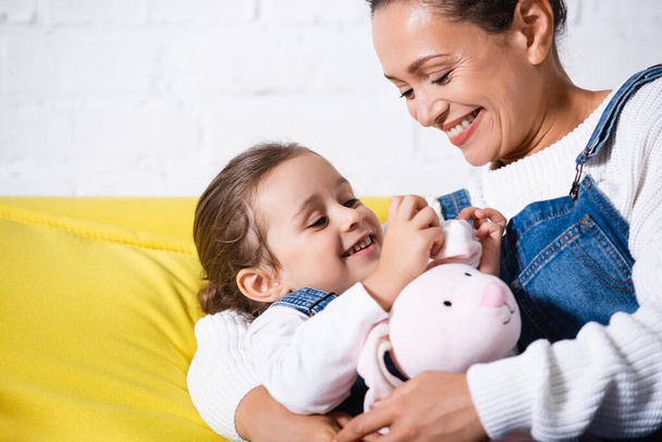 Mom hugging daughter with soft toy on yellow couch - Foto, Bild