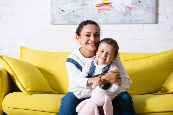 Mamma seduta sul divano e abbracciare la figlia con peluche a casa - Foto, immagini