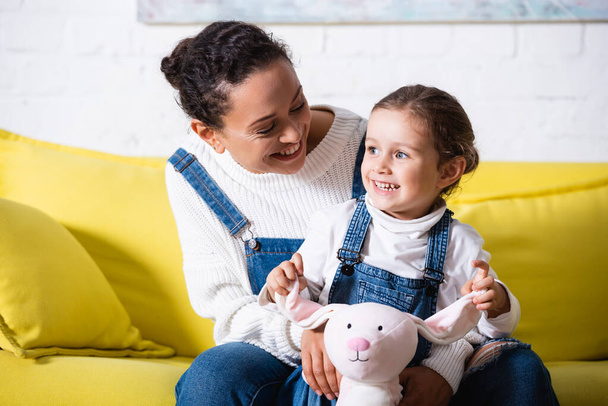 Mom sitting on couch and hugging daughter with soft toy at home - Foto, Bild