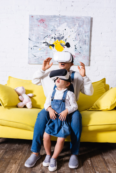 Emocionados madre e hija usando auriculares de realidad virtual cerca de sofá amarillo - Foto, imagen