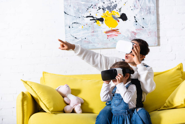 Mother pointing with finger and daughter using virtual reality headsets at home - Foto, imagen