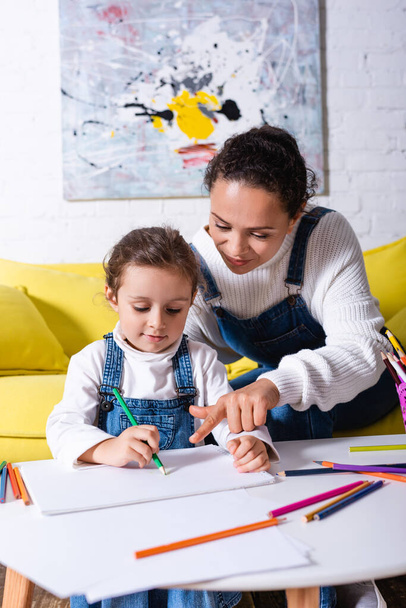 Mother pointing with finger and daughter drawing with color pencils at home - Φωτογραφία, εικόνα
