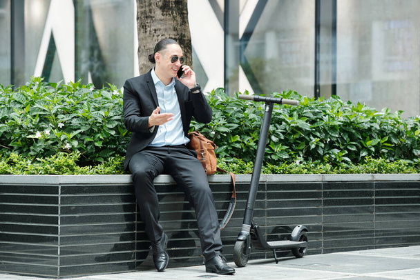 Lächelnder junger Geschäftsmann mit Sonnenbrille sitzt draußen neben seinem Motorroller und telefoniert mit einem Geschäftspartner - Foto, Bild