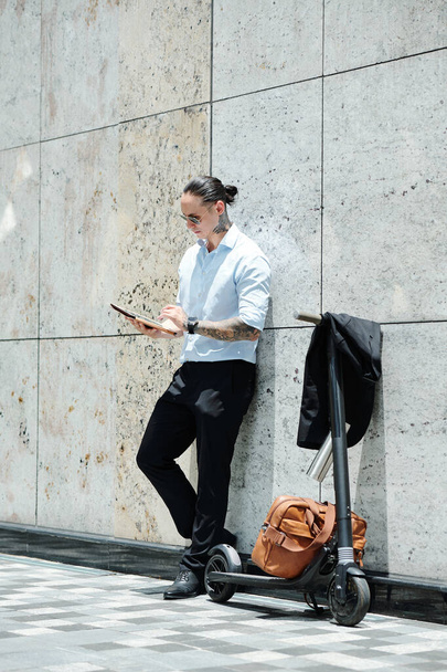Jeune homme d'affaires à la mode debout à l'extérieur à côté de son scooter et le document de lecture sur tablette numérique - Photo, image