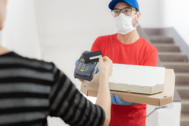 Delivery guy with protective mask holding pizza box and contactelss credit card terminal at the door. - Foto, Bild