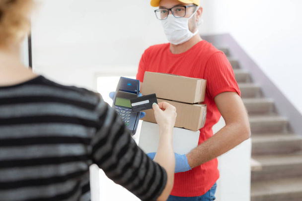 Delivery guy with a medical mask with packages and woman paying with contactless credit card at home during virus pandemic. - Photo, Image