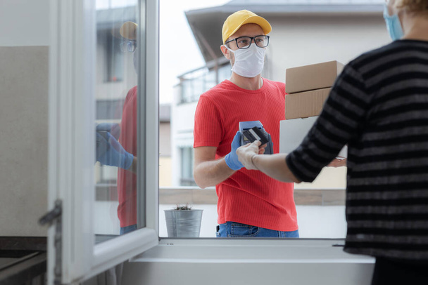Delivery guy with a medical mask with packages and woman paying with contactless credit card - delivering through a home window during virus pandemic. - Fotografie, Obrázek