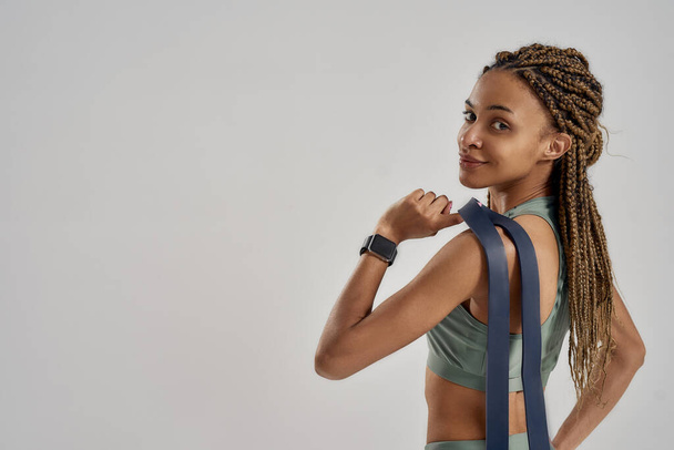 Staying healthy and fit. Happy young mixed race woman holding elastic band or fitness gum and smiling at camera while standing isolated over grey background, rear view - Foto, Bild