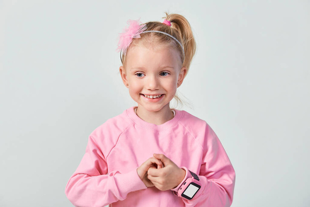 Cute caucasian little girl 4-5 year old wearing a smart watch on her wrist. Technology for children, against gray studio background - 写真・画像