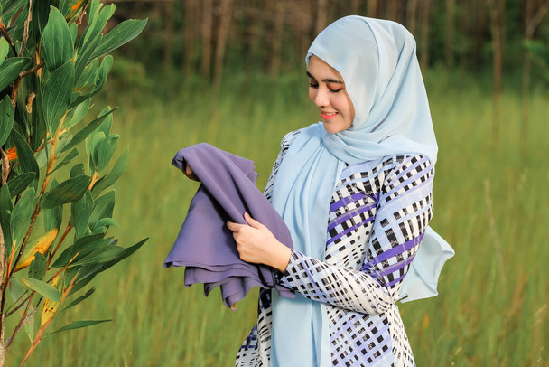 Portret van gelukkig mooi jong model in modieuze hijab stijl poseren in groen gebied terwijl het houden van stof. Stijlvolle moslim vrouwelijke hijab mode levensstijl portretteren concept. - Foto, afbeelding