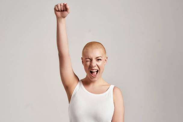 Portrait of a beautiful young caucasian woman with shaved head wearing white shirt, winking at camera and raising clenched fist, standing isolated over grey background - Фото, изображение