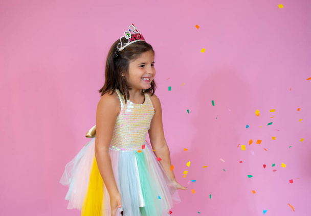 Girl in a festive dress stands on the pink background with confetti and smiles - Photo, Image