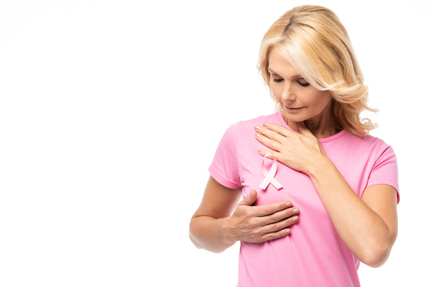 Ξανθιά γυναίκα αγγίζει το στήθος και κοιτάζοντας ροζ κορδέλα ευαισθητοποίησης του καρκίνου που απομονώνονται σε λευκό - Φωτογραφία, εικόνα