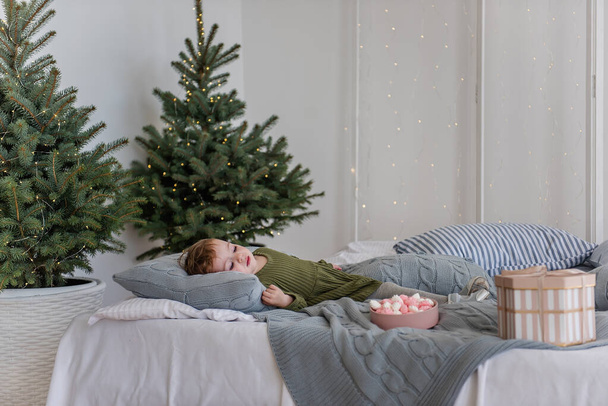 A little girl in a warm green dress lies on a white bed, among gray knitted pillows, eating a pink and white meringue. On a gray background garlands with lights, Christmas trees. Copy space, blurred - Valokuva, kuva