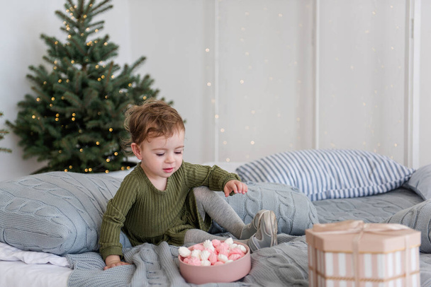 A little girl in a warm green dress lies on a white bed, among gray knitted pillows, eating a pink and white meringue. On a gray background garlands with lights, Christmas trees. Copy space, blurred - Fotografie, Obrázek
