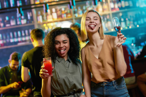 Attractive young women getting drunk, showing tongue to camera, posing with cocktail in their hands. Friends celebrating, having fun in the bar - Foto, Bild