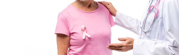 Panoramische opname van arts wijzend met de hand en knuffelende senior vrouw met roze lint op t-shirt geïsoleerd op wit - Foto, afbeelding
