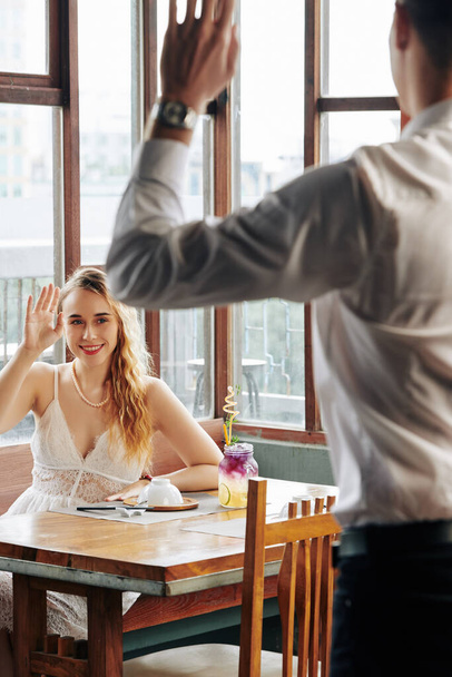 Aantrekkelijke jonge blonde vrouw zitten aan cafe tafel, glimlachen en zwaaien naar haar vriend - Foto, afbeelding