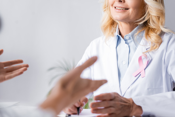 Selective focus of doctor in white coat with pink ribbon sitting near patient - Photo, image