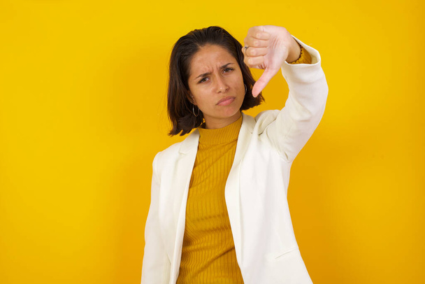 Discontent European woman shows disapproval sign, keeps thumb down, expresses dislike, frowns face in discontent, dressed in white jacket - Photo, Image