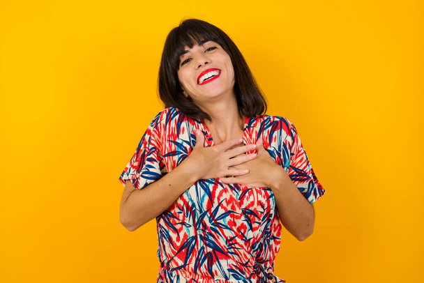 Happy woman standing and smiling on studio background has hands on chest near heart. Young emotional woman. Human emotions, facial expression concept. Front view. - Фото, изображение