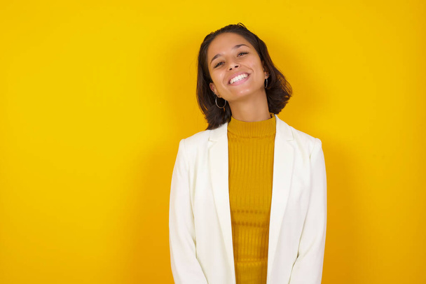 Beautiful smiling brunette businesswoman standing against yellow background with broad smile, shows white teeth, wears casual t shirt, stands over studio wall rejoices having day off. Woman standing indoors. - 写真・画像