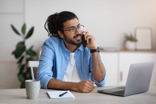 Millennial Indian Mann spricht mit Handy und Laptop im Home Office - Foto, Bild