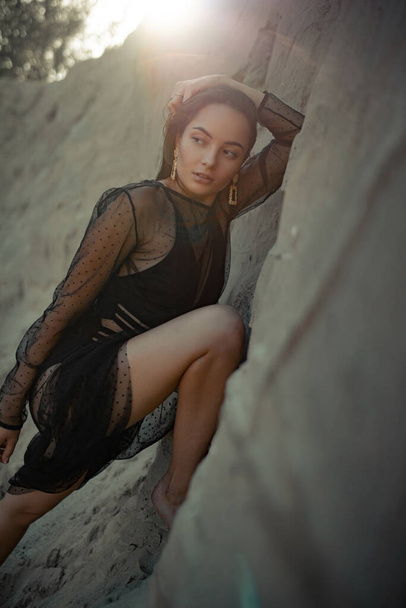 Hübsche Frau im schwarzen transparenten Kleid posiert in einer Sandschlucht. Gegenlicht. - Foto, Bild