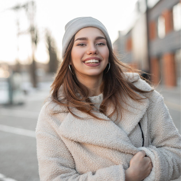Portrait of a happy attractive young woman with a beautiful smile in a trendy faux fur coat in a knitted stylish hat on the street in the city. Cute girl posing and smiling outdoors on a spring day. - Foto, afbeelding