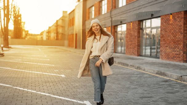 Joyful young woman in fashionable clothes is walking in the parking on background a bright sunset. Happy girl in trendy outerwear with a positive smile enjoys a bright sun - Foto, Bild