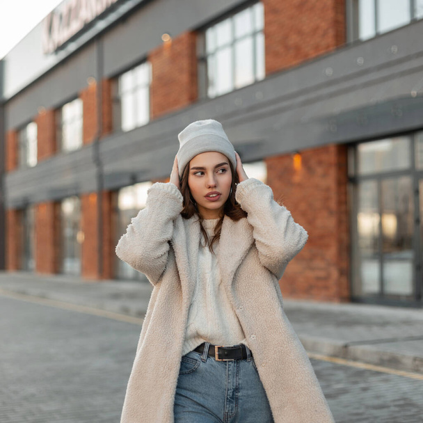 Trendy urban young woman straightens stylish gray knitted hat outdoors. Attractive girl model in a fashionable faux fur coat in blue jeans posing on the road near a vintage building in the street. - Foto, Bild
