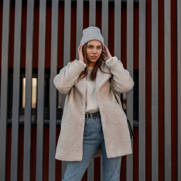 ヴィンテージブルーのスタイリッシュなジーンズのスタイリッシュなエココートのかなりファッショナブルな若い女性は、都市の近代的な金属壁の近くにトレンディーなグレーのニット帽をまっすぐにします。 - 写真・画像