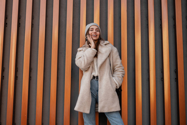 ブルージーンズのトレンディーなエコファーコートを着た都会の若い女性が、革のハンドバッグを持ったセーターの灰色の帽子を編んで、路上で積極的に屋外で笑顔を見せます。金属の壁の近くのかわいい女の子. - 写真・画像